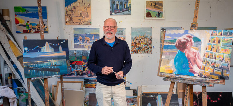 man holding paint brush in art studio, surrounded by paintings 