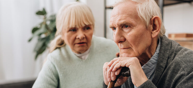 elderly man who looks troubled with head on hands atop cane and woman beside him, looks concerned