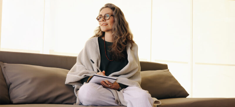 older adult woman holding pen and paper seated on a bed