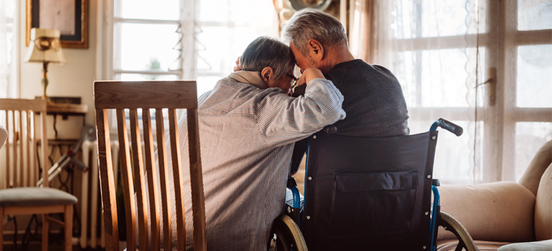 two elderly men seated (one in a wheelchair) leaning on each other