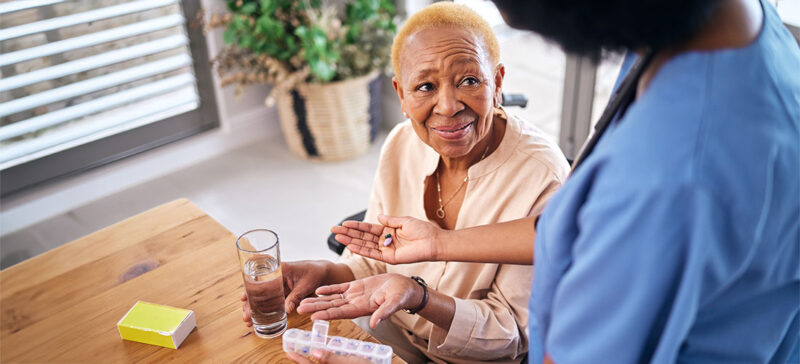 elderly woman with weekly pill box and glass of water nearby holding her hand open for healthcare worker with medication in hand