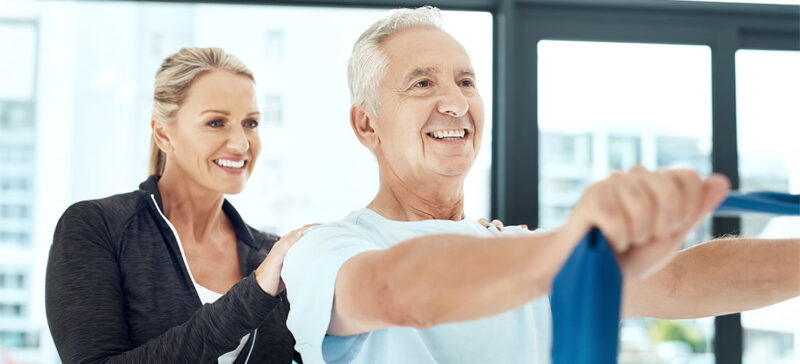 older gentleman on exercise equipment with younger person encouraging from behind