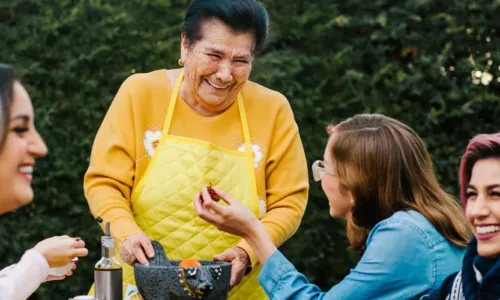 a group of older adults gathered around a table outside with one standing, and one younger person seated