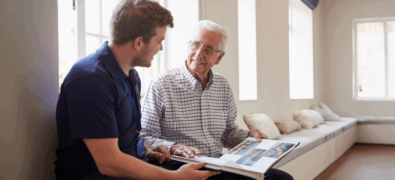 older adult man seated next to younger man, looking at photo album and smiling at eachother
