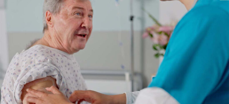 smiling, older adult gentleman in a hospital gown seated facing a medical professional who is preparing him for a vaccine